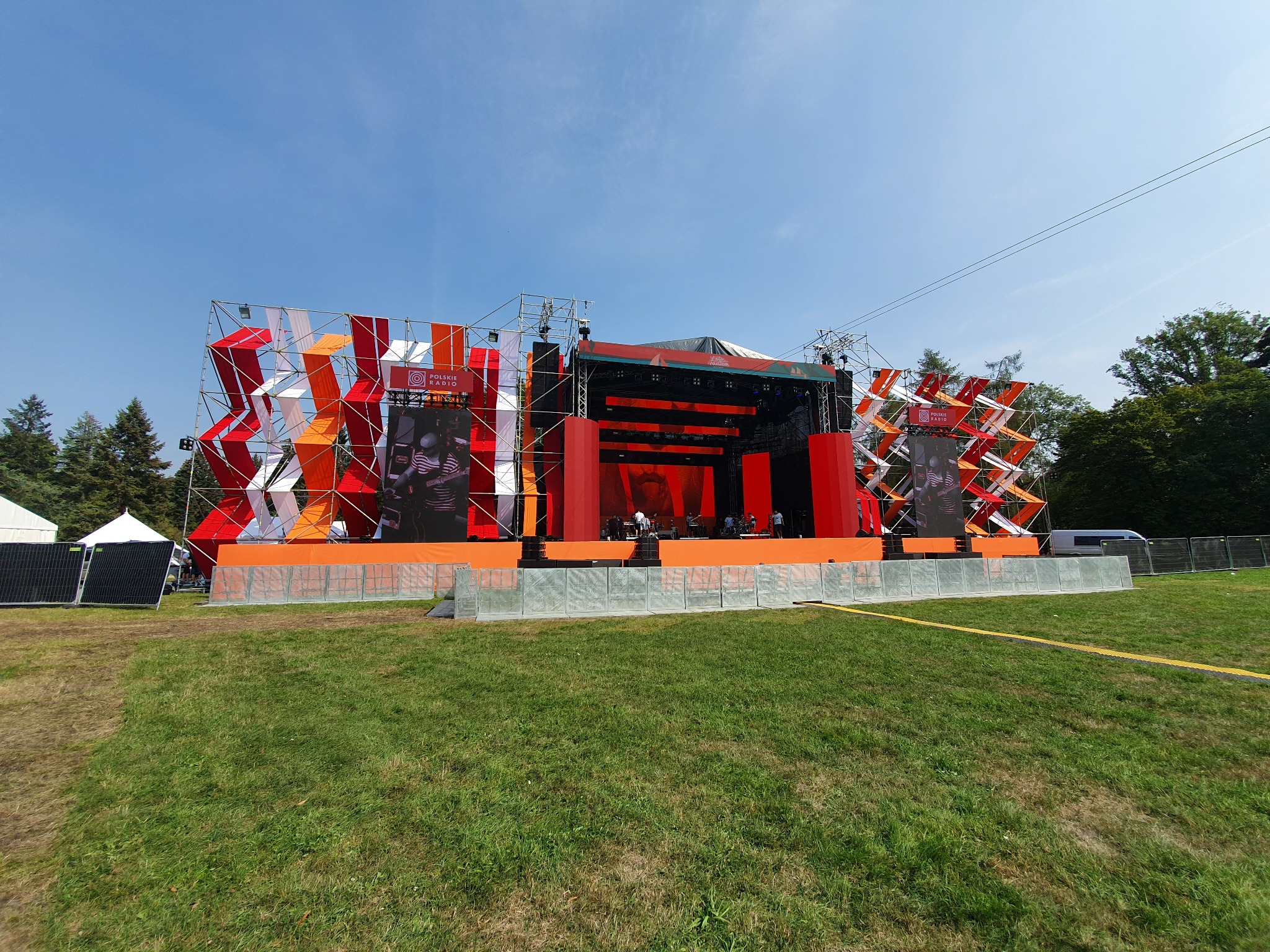Szeroki kadr na scenę plenerową z metalową konstrukcją ozdobioną czerwono-białymi elementami, z widocznymi ekranami LED i zespołem przygotowującym się do występu, na tle zielonej trawy i błękitnego...