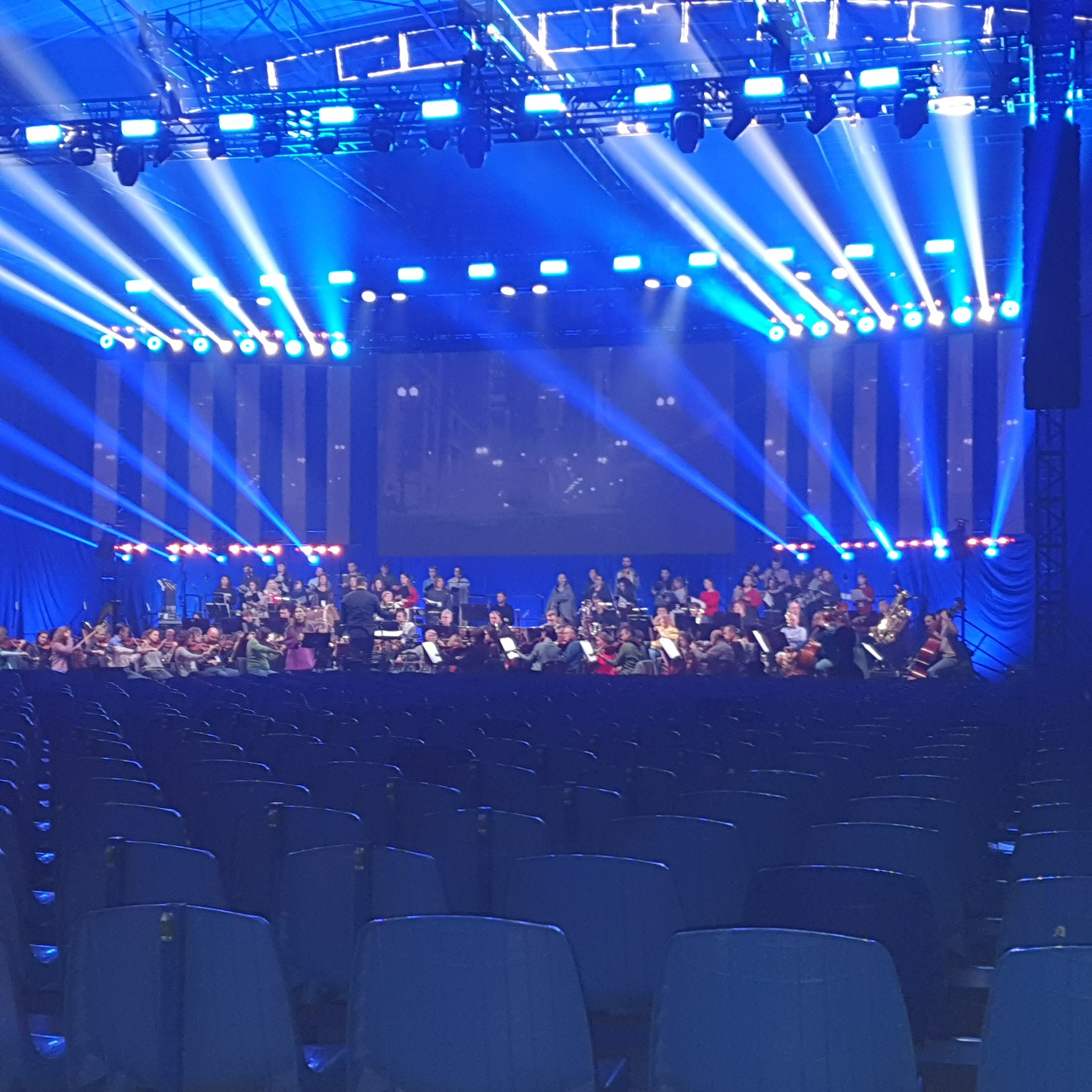 Orkiestra symfoniczna na scenie oświetlonej niebieskimi reflektorami, widok z pustych rzędów krzeseł.