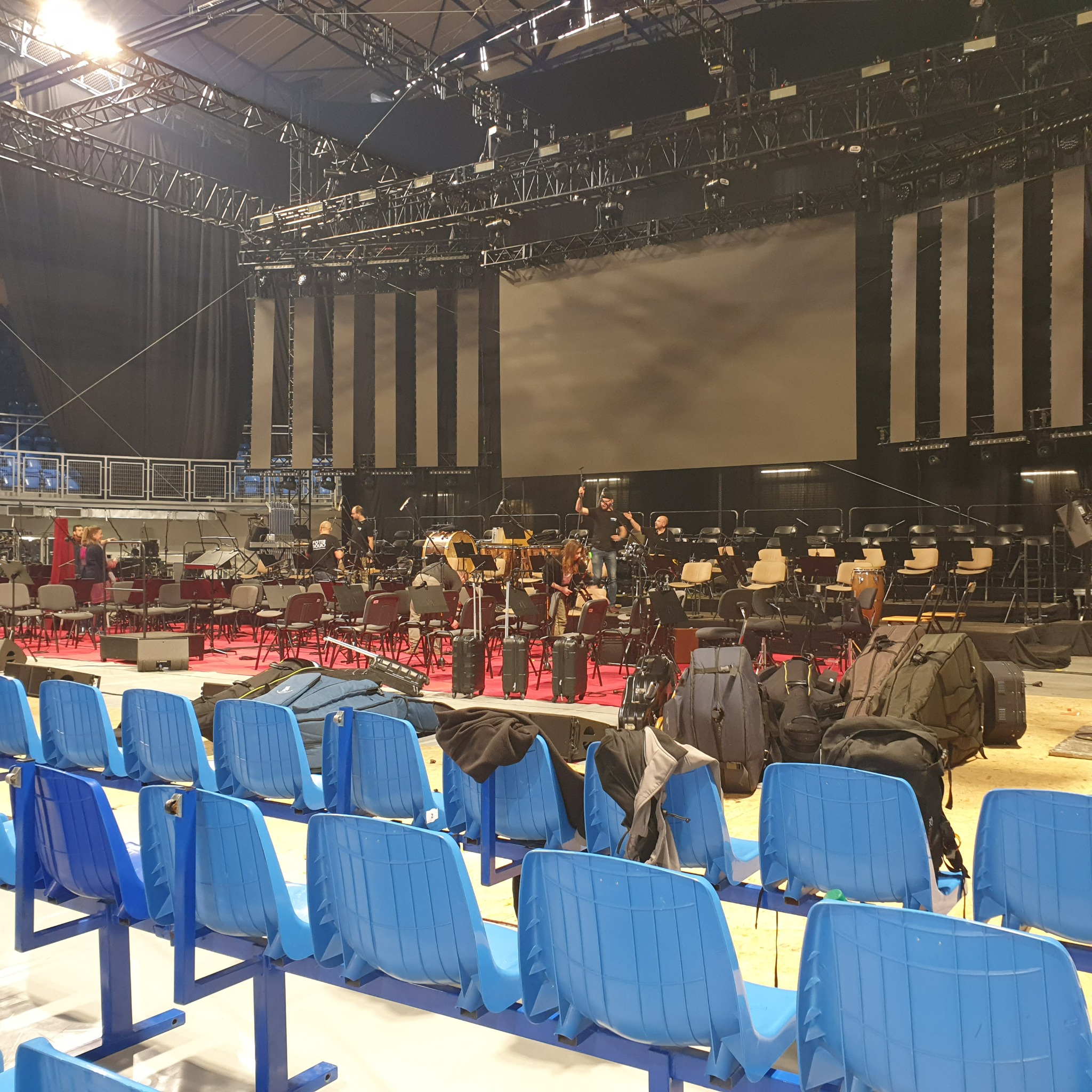 Przygotowania do koncertu: scena z instrumentami, krzesłami dla muzyków i publiczności, bagaże oraz ekipa techniczna w trakcie rozkładania sprzętu.