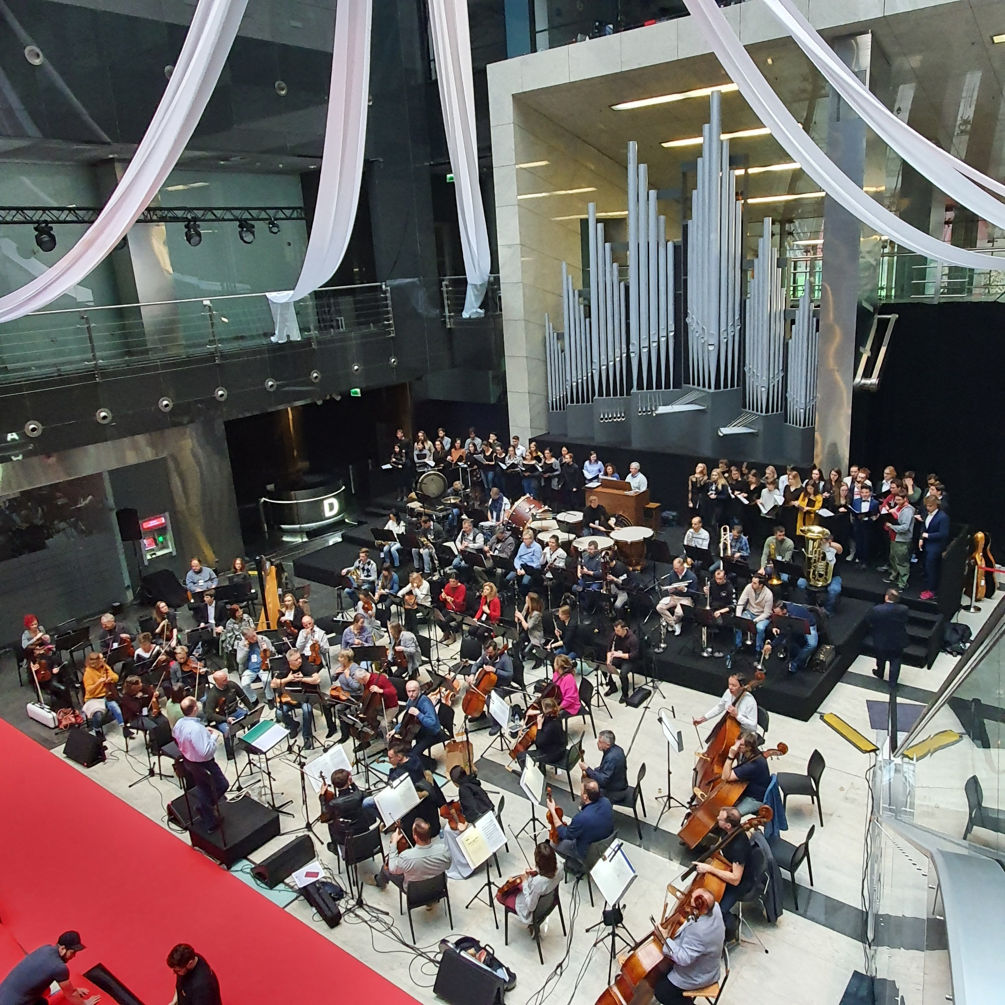 Widok z góry na orkiestrę symfoniczną i chór podczas występu w nowoczesnym wnętrzu z organami piszczałkowymi w tle, udekorowanym białymi draperiami.