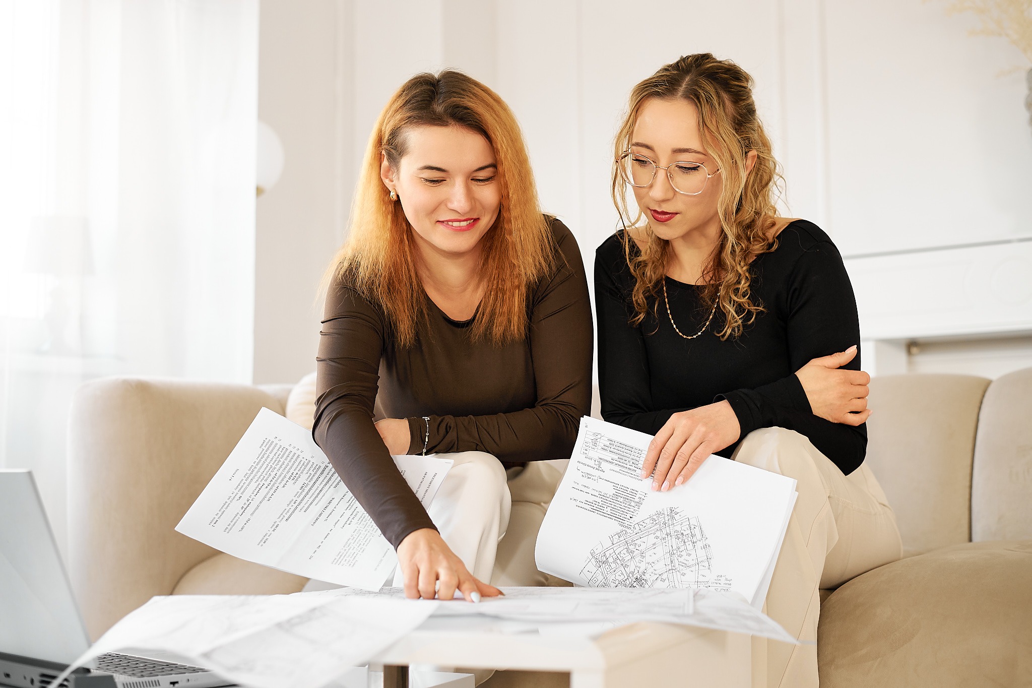 Dwie kobiety analizują plany architektoniczne na papierze, siedząc na beżowej sofie z laptopem w tle, w jasnym wnętrzu.