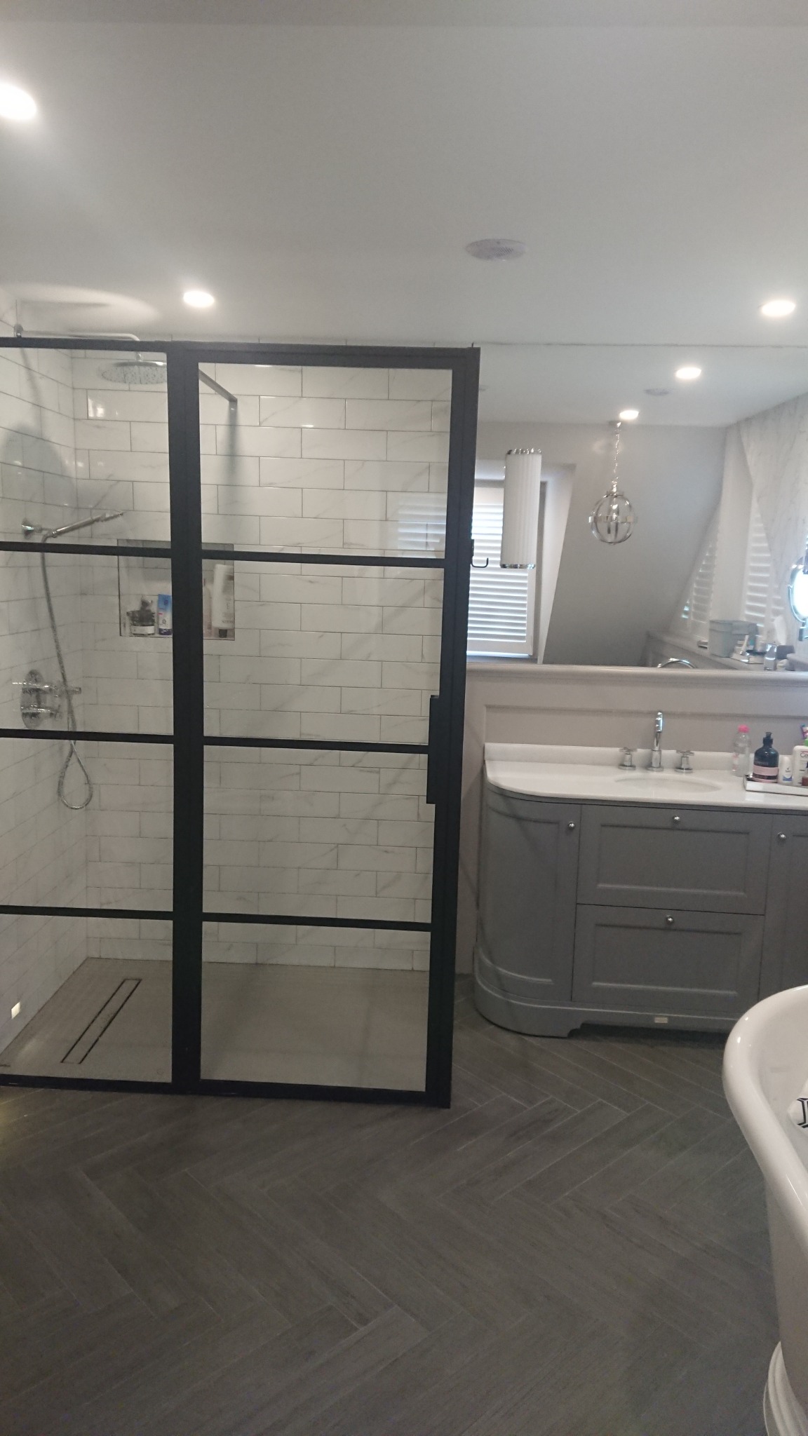 Nowoczesna łazienka z kabiną prysznicową typu walk-in z czarnymi profilami, szarą szafką pod umywalką i podłogą ułożoną w jodełkę.