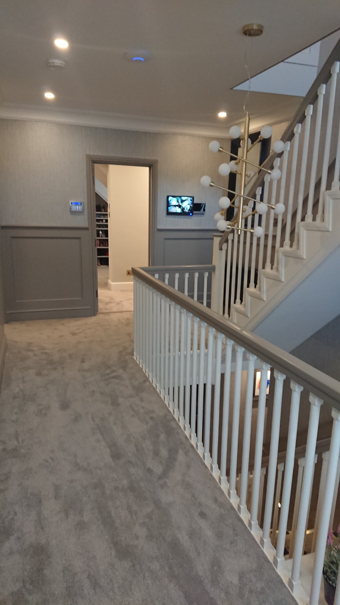 Nowoczesny korytarz z szarą wykładziną, białymi schodami z szarą balustradą, złotą lampą wiszącą i telewizorem na ścianie.