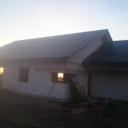 Dom jednorodzinny w trakcie budowy z częściowo położonym, ciemnym pokryciem dachowym na tle zachodzącego słońca.