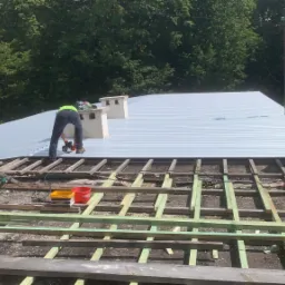 Dwóch dekarzy montuje blachodachówkę na dachu domu, widoczny szkielet dachu z łat i kontrłat, w tle drzewa i niebo.