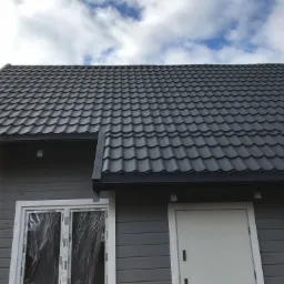 Szary dom z blachodachówką, okno zabezpieczone folią ochronną, widoczne błękitne niebo z chmurami.