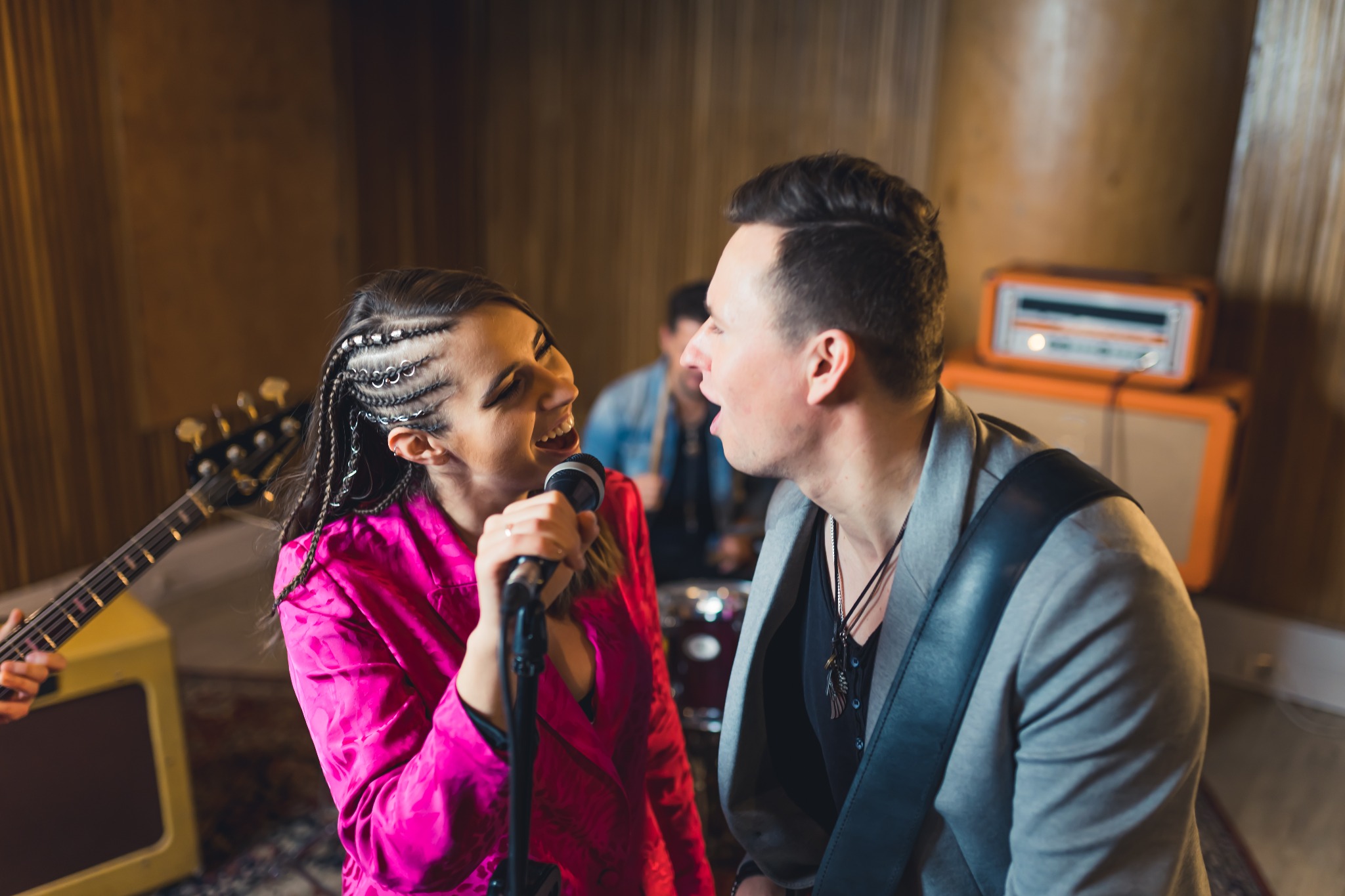 Wokalistka w różowej koszuli śpiewa do mikrofonu, obok wokalista z gitarą i perkusista w tle, podczas występu na scenie.