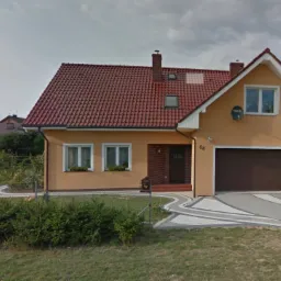 Dwupiętrowy dom z garażem, pomalowany na pomarańczowo, z brązową bramą garażową i czerwoną dachówką, widoczny również zadbany ogród z kwiatami i krzewami.