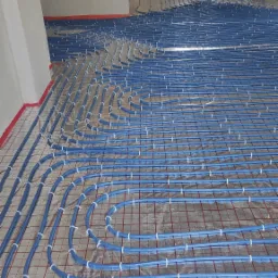 Niebieskie rury ogrzewania podłogowego ułożone na folii izolacyjnej z czerwoną siatką, widoczne w surowym wnętrzu pomieszczenia w trakcie instalacji.