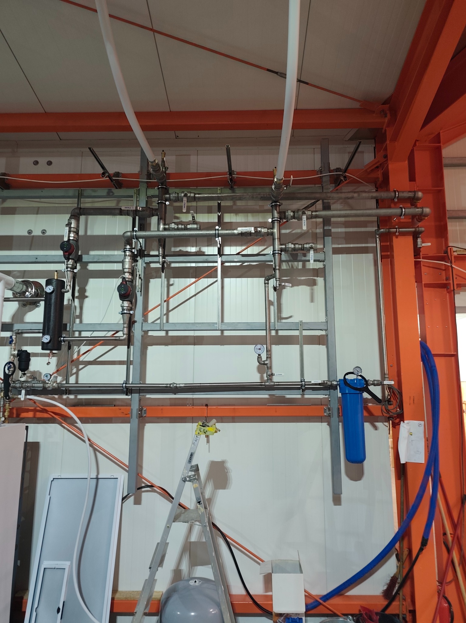 Kompleksowa instalacja hydrauliczna z filtrem wody, rurami i zaworami, zamontowana na ścianie w pomieszczeniu przemysłowym. Widoczna drabina i elementy konstrukcyjne.