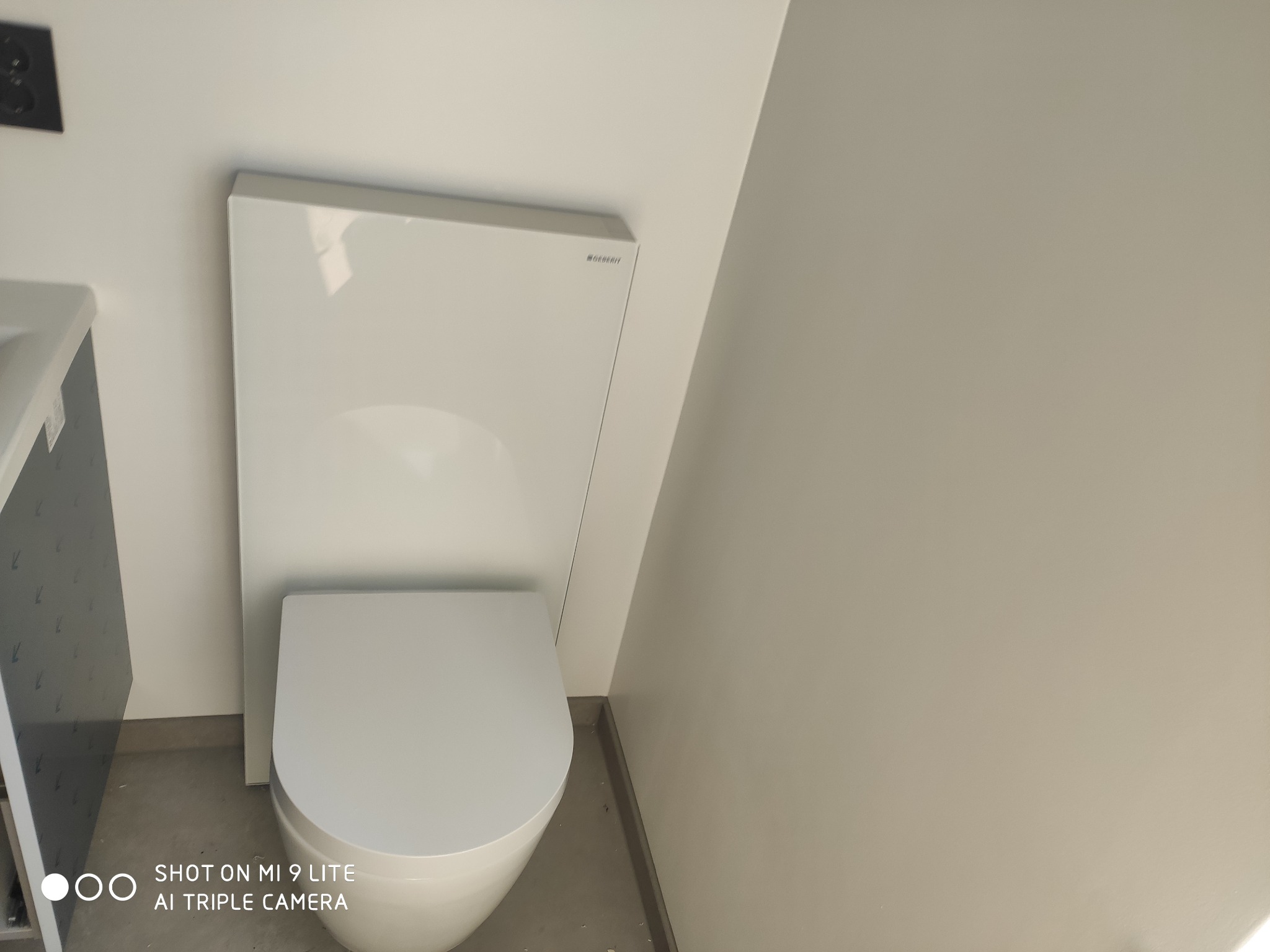 Nowoczesna, podwieszana toaleta Geberit z białą obudową, widoczna instalacja podtynkowa, minimalistyczne wnętrze łazienki w odcieniach bieli i szarości.