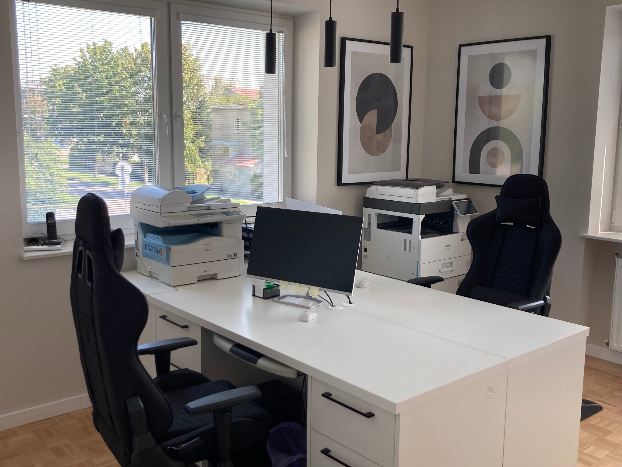 Jasne biuro z białym biurkiem, dwoma drukarkami, monitorem i dwoma czarnymi fotelami biurowymi. Na ścianie wiszą abstrakcyjne obrazy w czarnych ramkach.