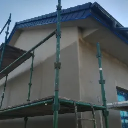 Budynek w trakcie prac elewacyjnych, widoczne rusztowanie, surowy tynk i zabezpieczone okno folią, ciemna blacha na dachu z rynną.