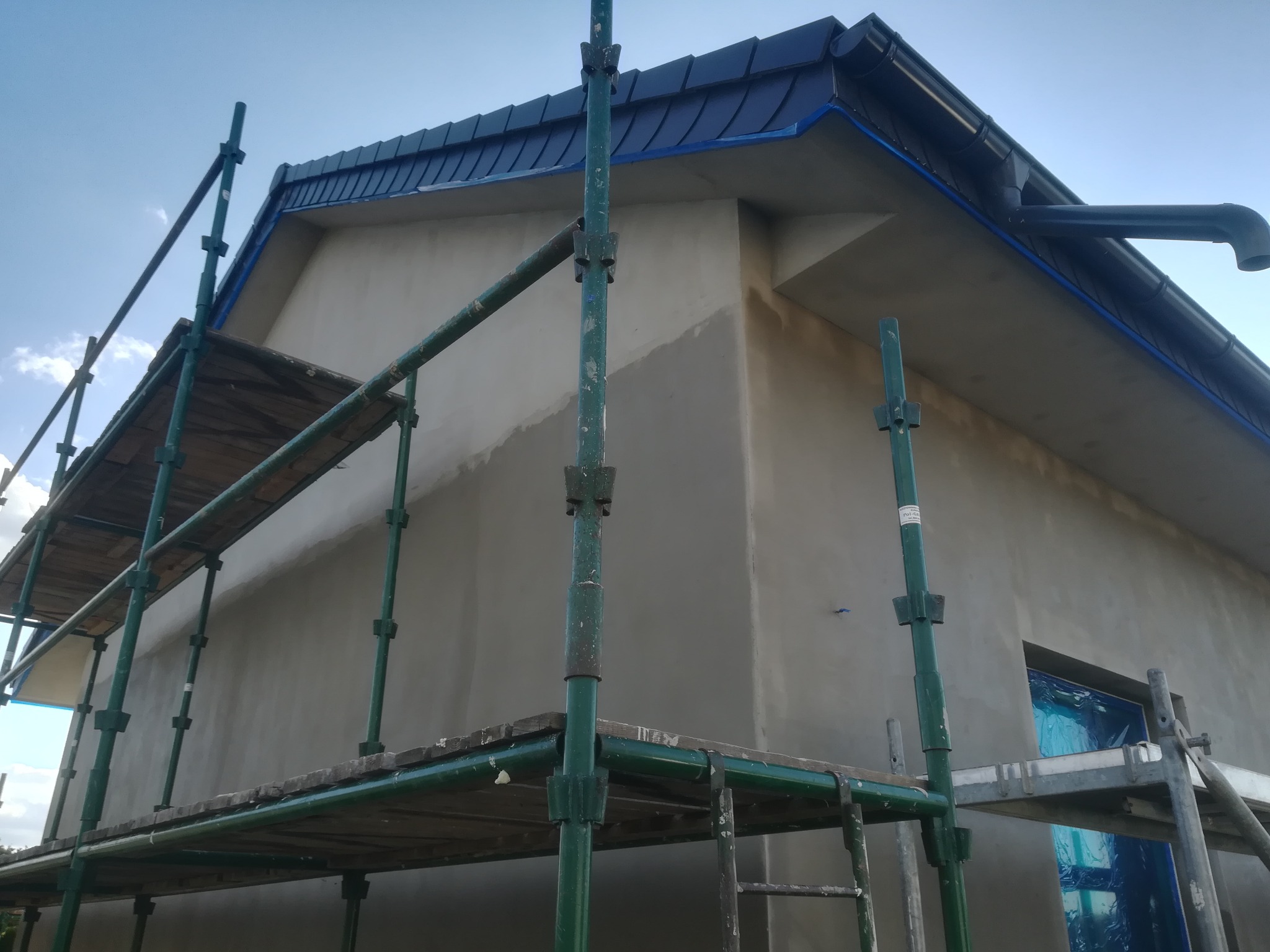 Budynek w trakcie prac elewacyjnych, widoczne rusztowanie, surowy tynk i zabezpieczone okno folią, ciemna blacha na dachu z rynną.