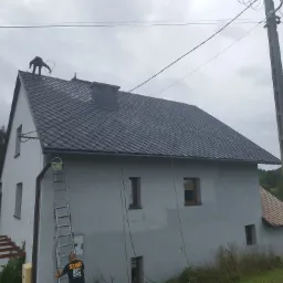 Pracownik na dachu domu jednorodzinnego z szarym dachem z dachówki ceramicznej, drabina oparta o ścianę, widoczny pomalowany komin i linia energetyczna.