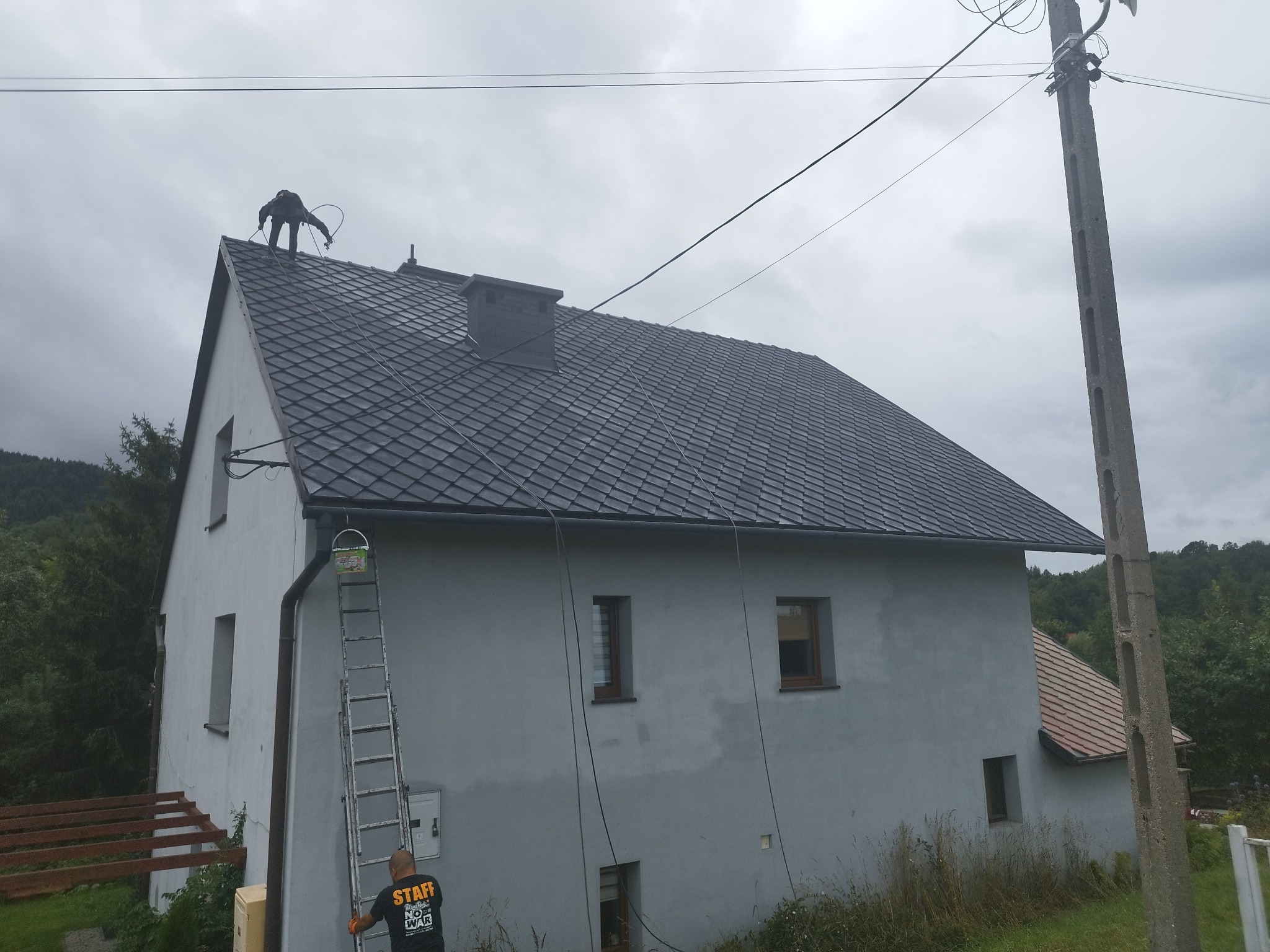 Pracownik na dachu domu jednorodzinnego z szarym dachem z dachówki ceramicznej, drabina oparta o ścianę, widoczny pomalowany komin i linia energetyczna.