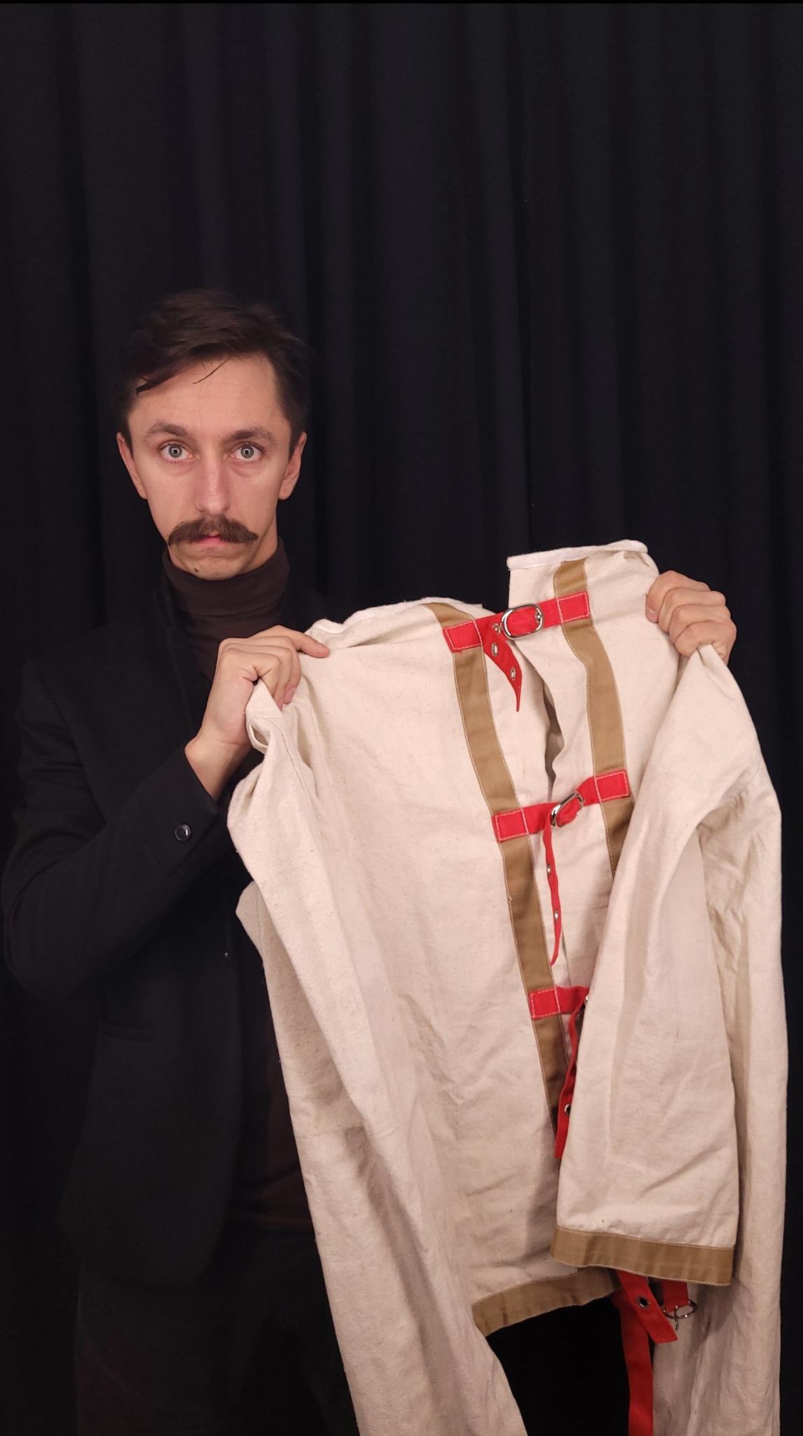 Mężczyzna z wąsami w czarnej marynarce prezentuje kaftan bezpieczeństwa z czerwonymi i brązowymi paskami na tle czarnej zasłony.