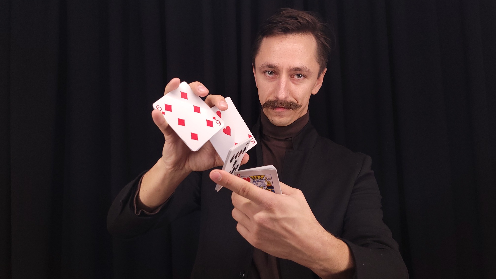 Mężczyzna z wąsami w czarnym garniturze prezentuje trik z kartami do gry na czarnym tle.
