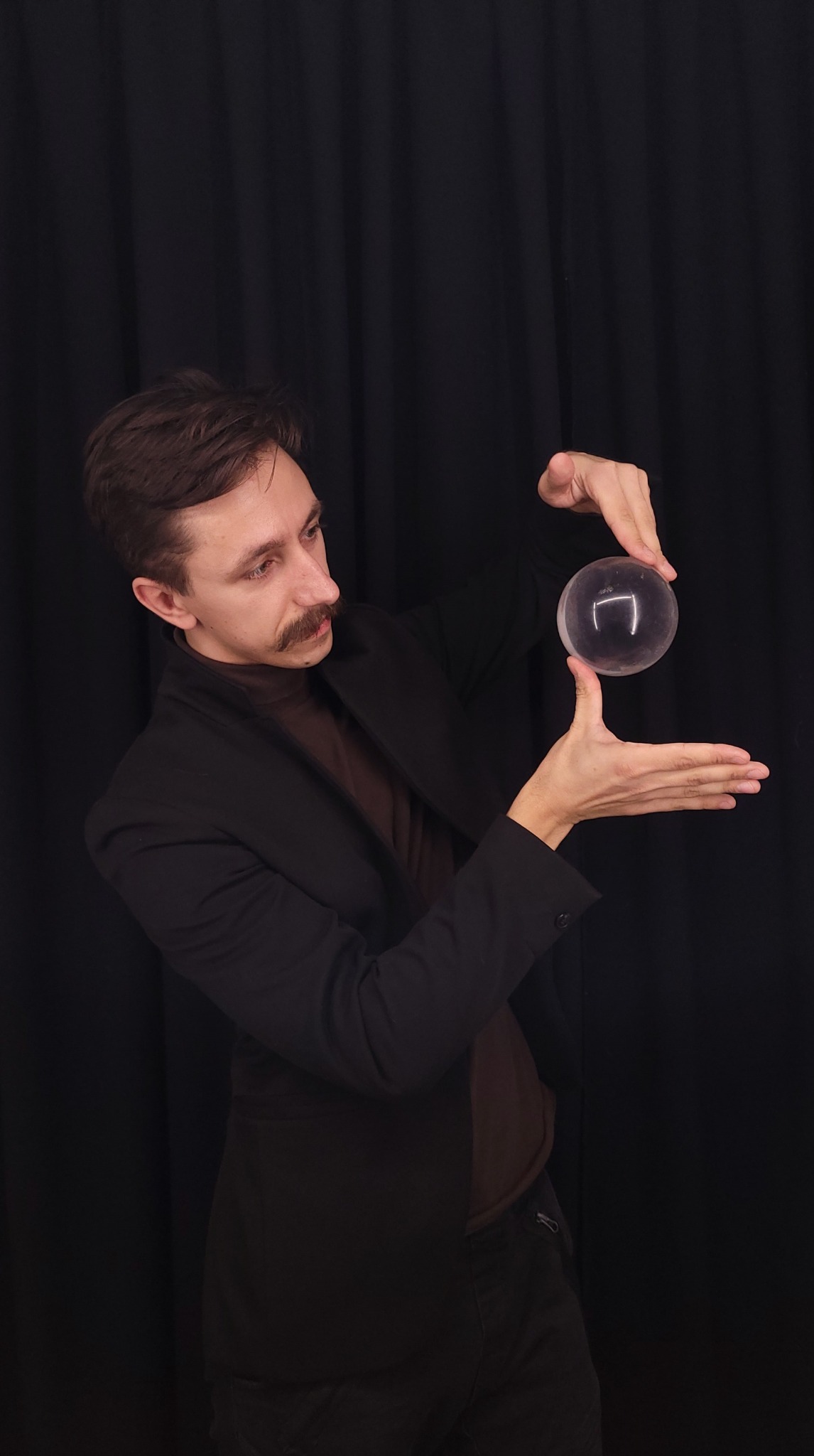 Mężczyzna z wąsami, w brązowym golfie i czarnej marynarce, prezentuje lewitującą, przezroczystą kulę na tle czarnej zasłony.