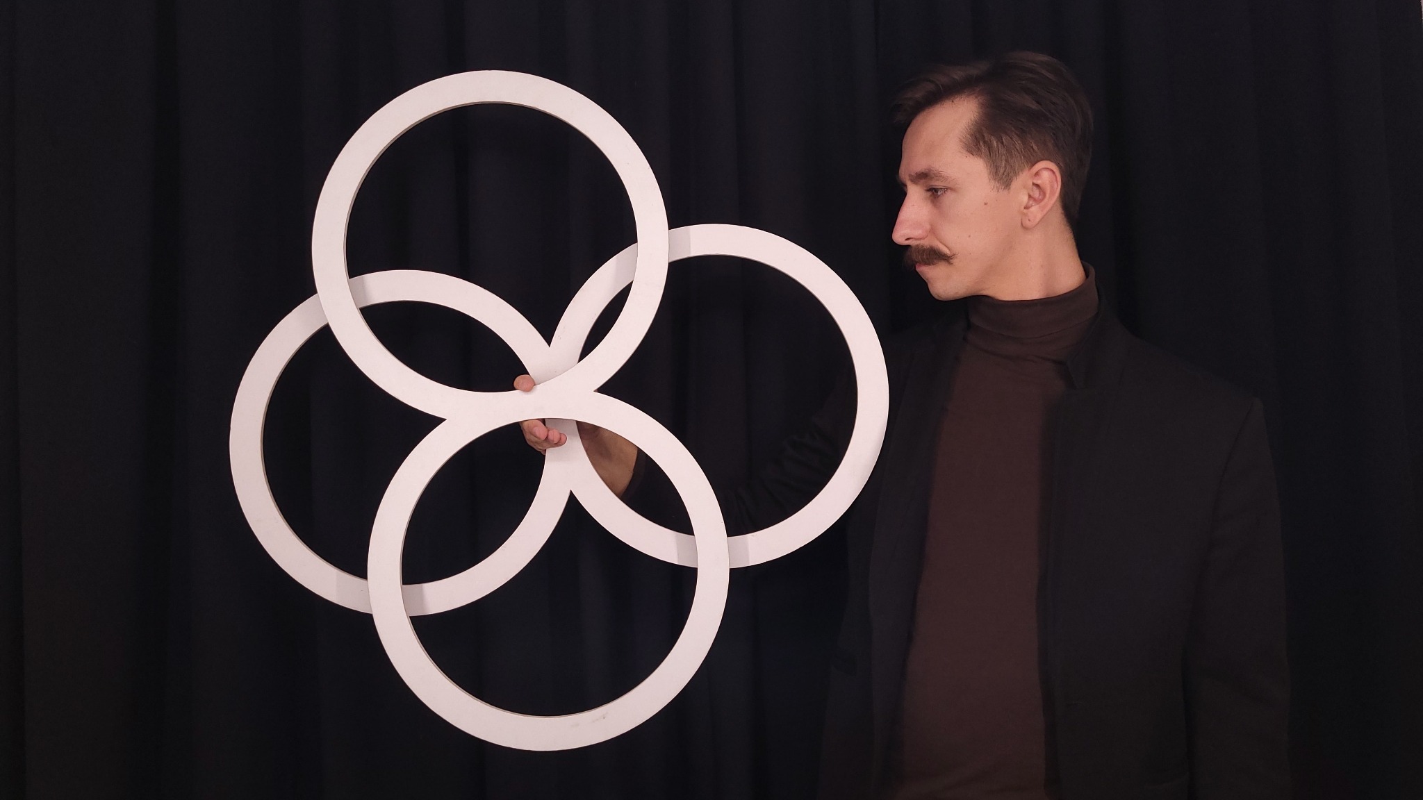 Mężczyzna z wąsami w czarnej marynarce prezentuje splecione białe okręgi na tle czarnej zasłony.