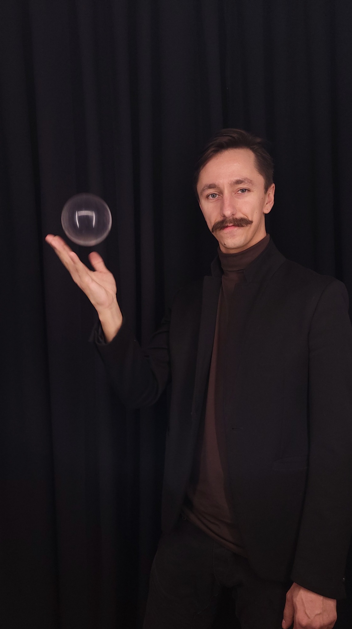 Mężczyzna z wąsami w czarnej marynarce unosi przezroczystą kulę na dłoni przed czarnym tłem.