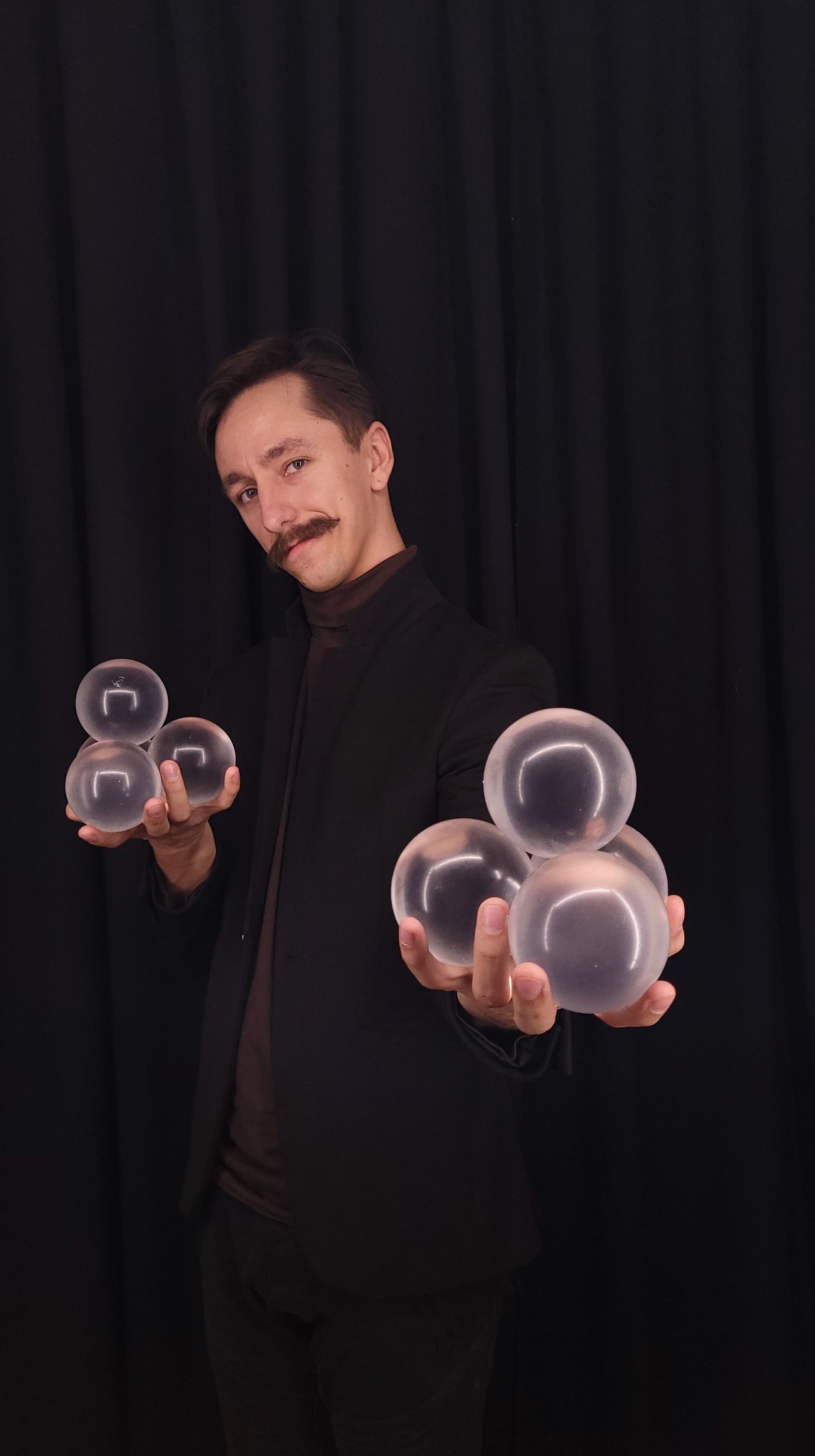 Iluzjonista z Rzeszowa prezentuje pokaz z przezroczystymi kulami na tle czarnej kurtyny. Uwagę zwracają charakterystyczne wąsy artysty.