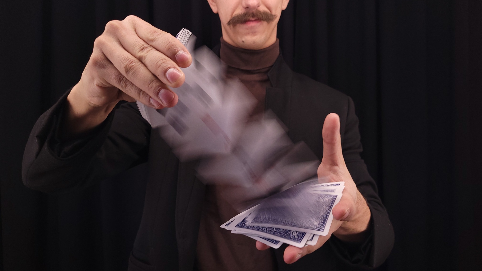 Iluzjonista w brązowym golfie i czarnej marynarce tasuje talię kart na czarnym tle, tworząc efekt rozmycia ruchu.
