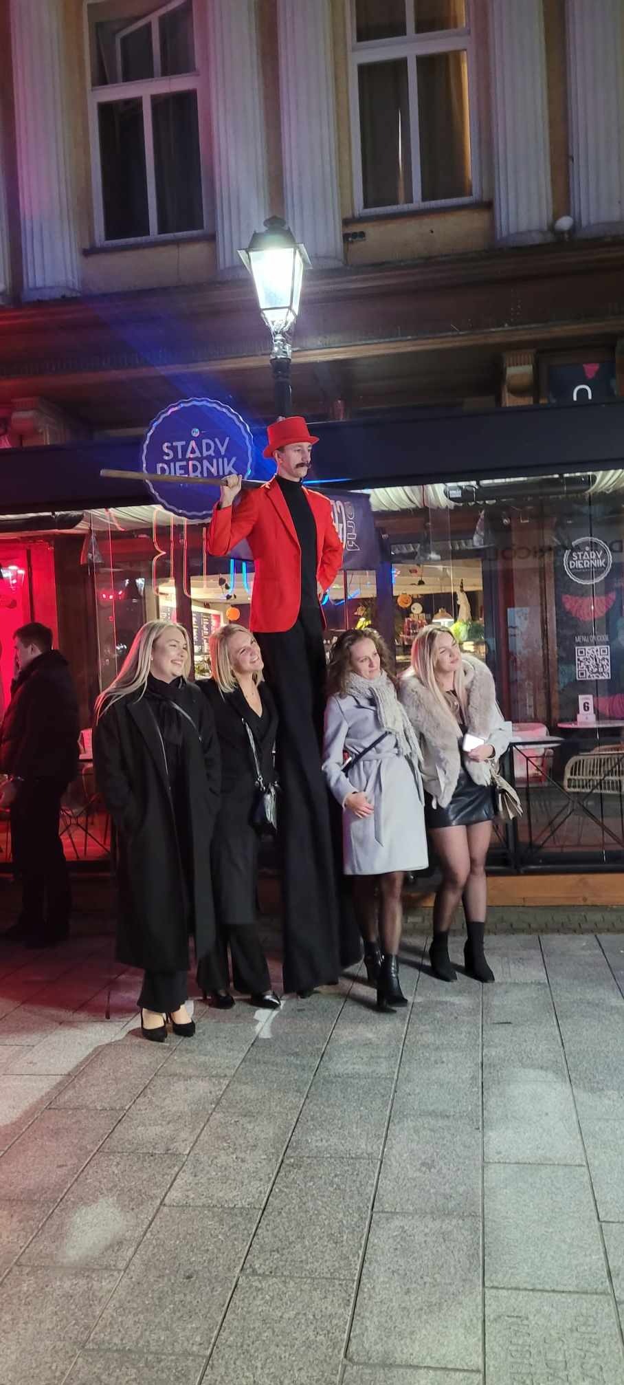 Iluzjonista na szczudłach w czerwonym fraku i kapeluszu pozuje do zdjęcia z grupą kobiet na tle lokalu 'Stary Dziennik'.
