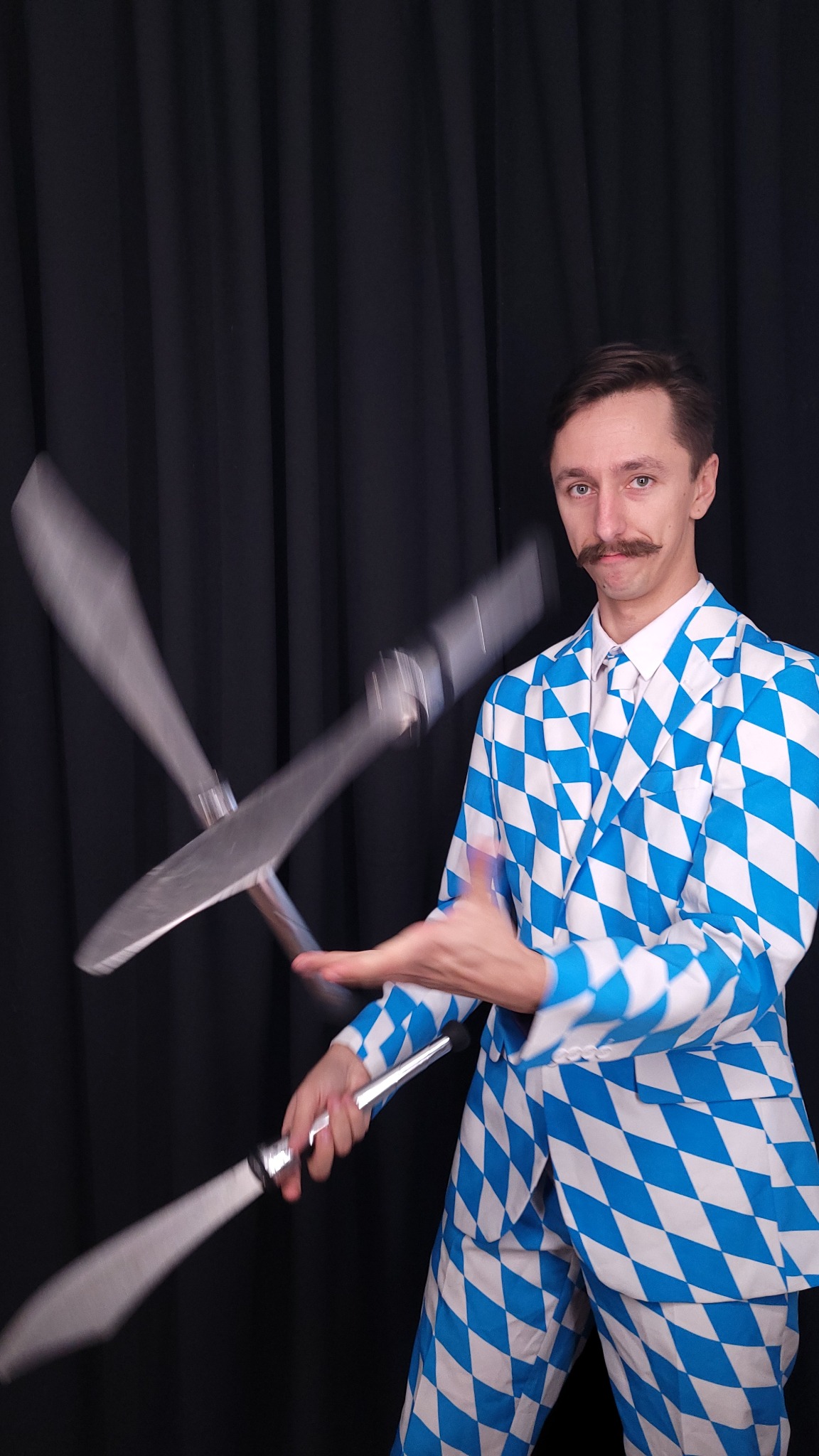Mężczyzna w niebiesko-białym kostiumie w romby żongluje srebrnymi maczugami na tle czarnej zasłony.