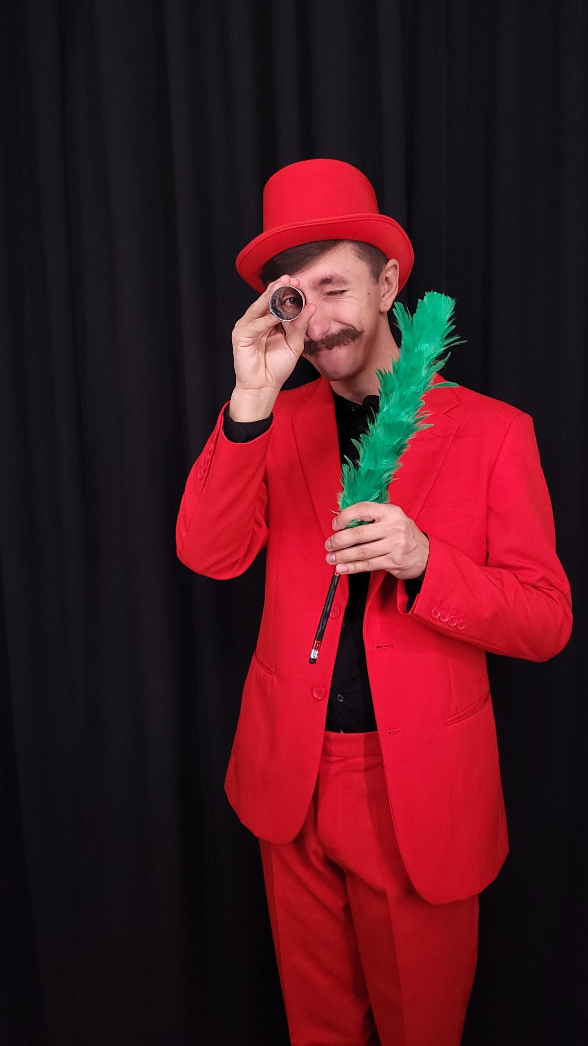 Mężczyzna z wąsami w czerwonym garniturze i kapeluszu patrzy przez metalową rurkę, trzymając zielone pióro na tle czarnej zasłony.
