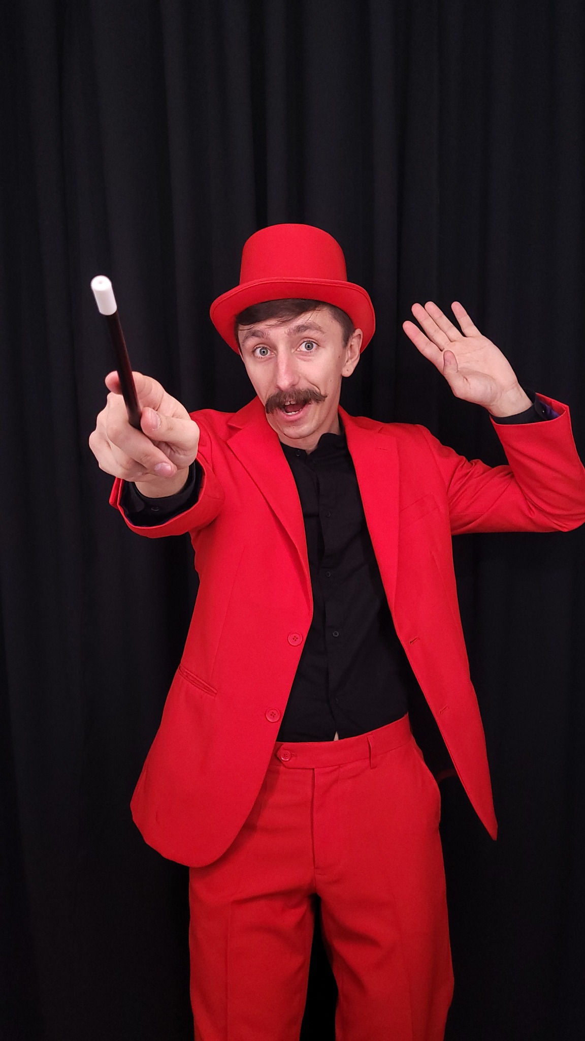 Mężczyzna z wąsami w czerwonym garniturze i kapeluszu, trzymający w dłoni różdżkę na tle czarnej kurtyny.