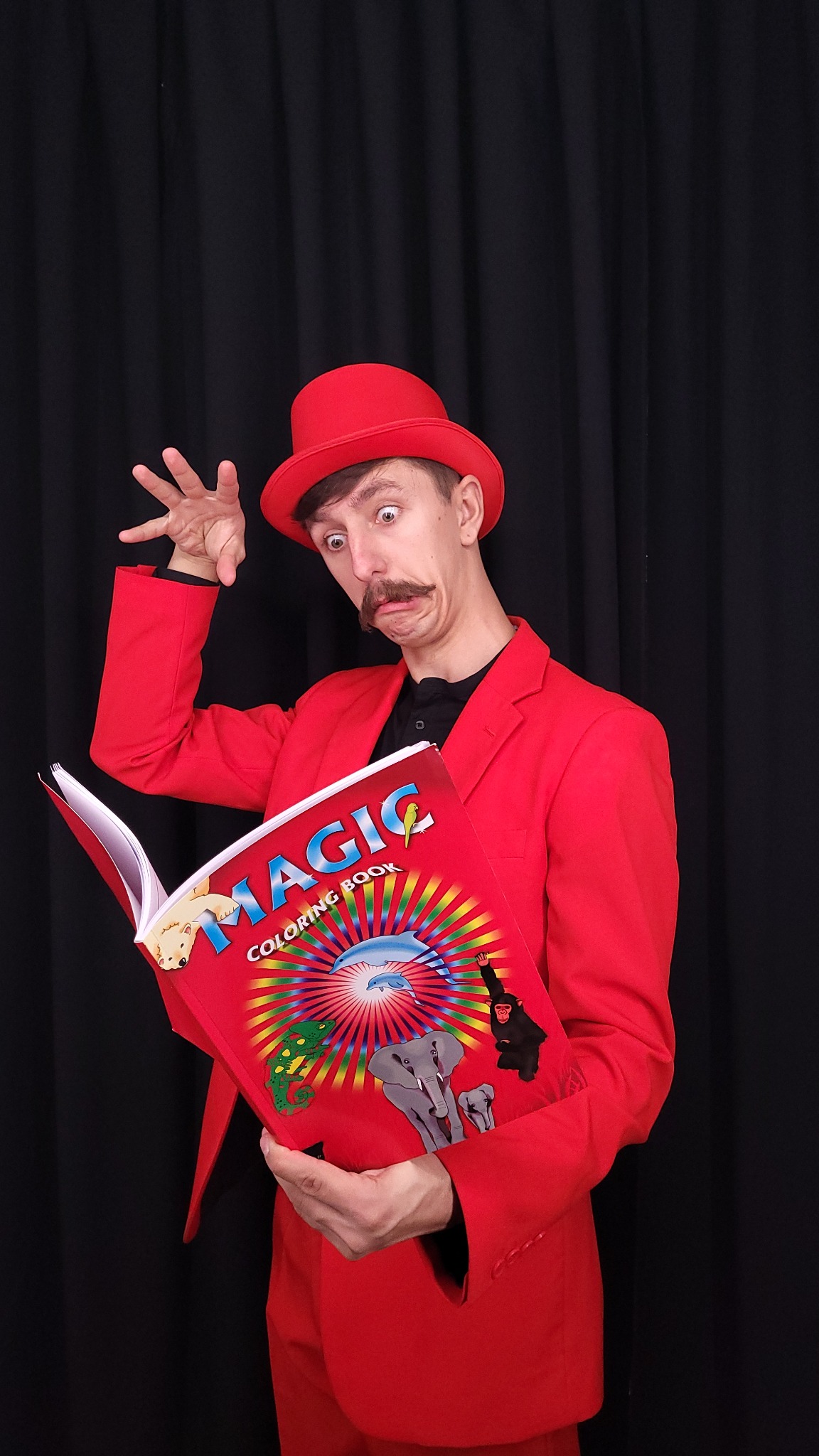 Iluzjonista w czerwonym garniturze i meloniku prezentuje 'Magic Coloring Book' na tle czarnej kotary, z wyrazem zaskoczenia na twarzy.