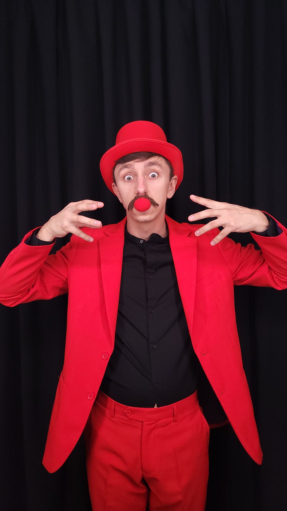 Mężczyzna w czerwonym garniturze i kapeluszu z czerwonym nosem klauna na tle czarnej zasłony, wykonujący gest dłońmi.