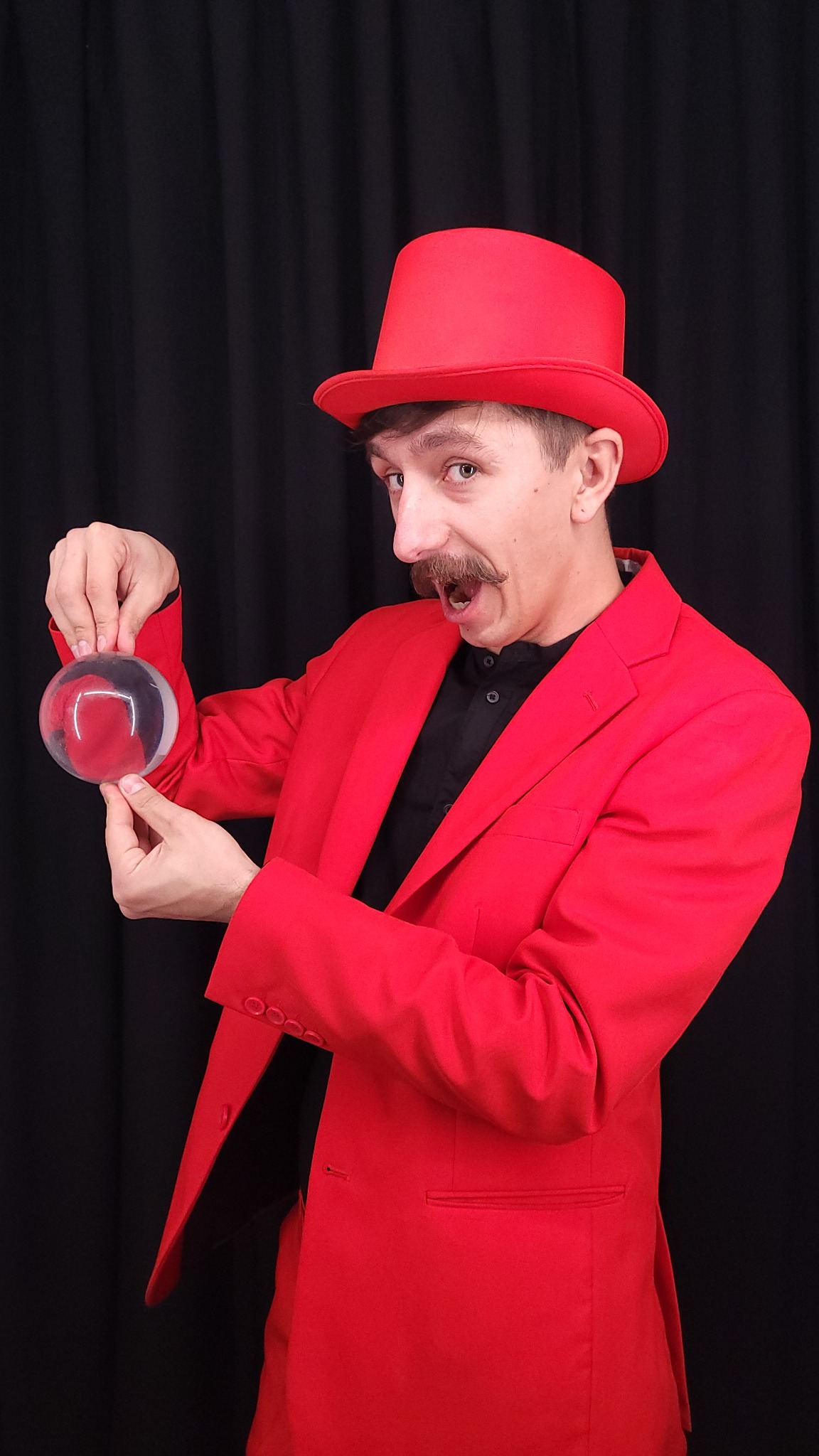 Mężczyzna w czerwonym fraku i cylindrze prezentuje przezroczystą kulę, z zaciekawioną miną na tle czarnych zasłon.