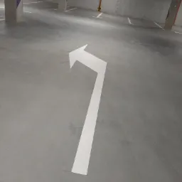 Strzałka kierunkowa na betonowej posadzce parkingu podziemnego, widoczne ślady opon, słupy z fioletowymi elementami, oświetlenie sufitowe.