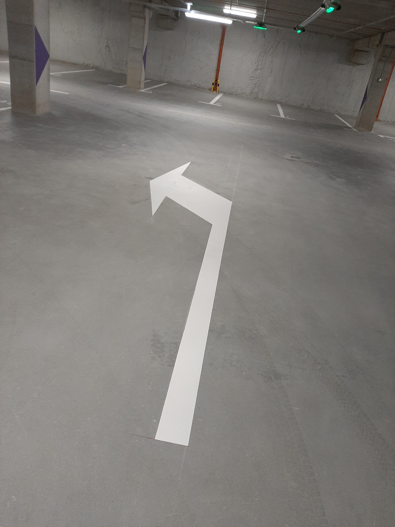 Strzałka kierunkowa na betonowej posadzce parkingu podziemnego, widoczne ślady opon, słupy z fioletowymi elementami, oświetlenie sufitowe.