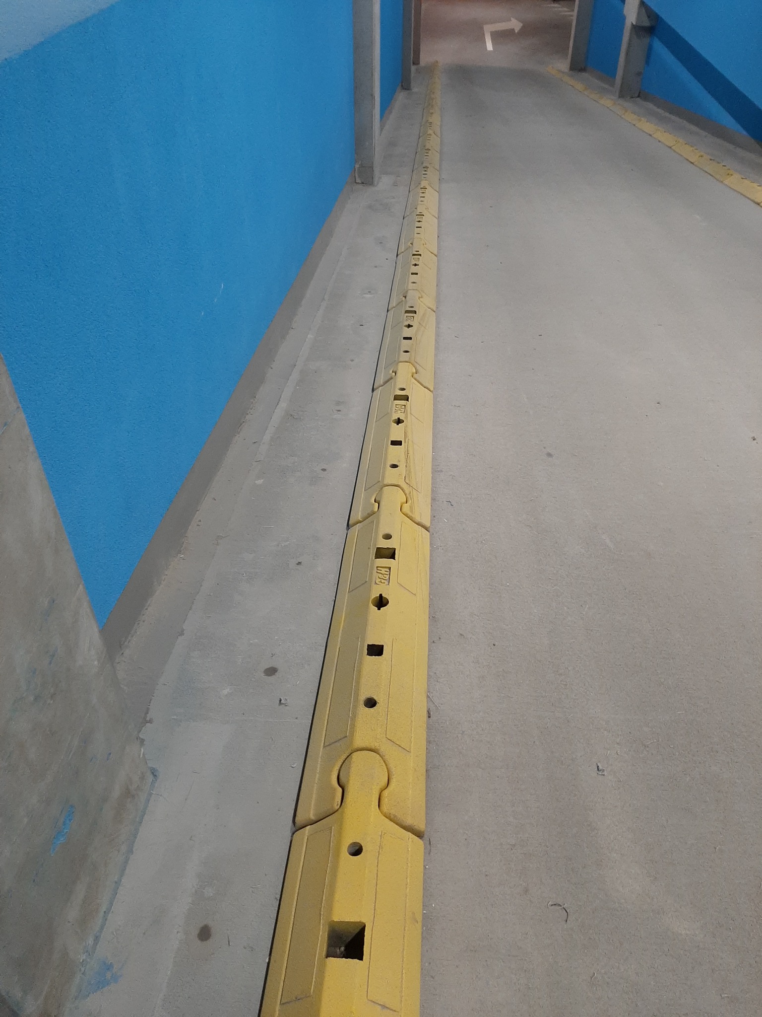 Żółta bariera ochronna zamontowana wzdłuż ściany w garażu podziemnym, wskazująca kierunek jazdy.