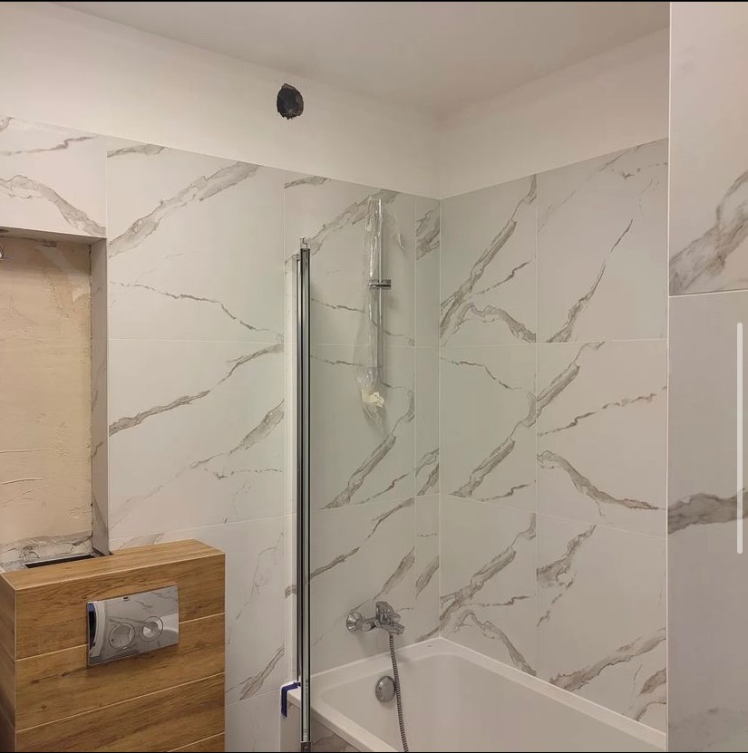 Wykończona łazienka z białymi płytkami imitującymi marmur, widoczna wanna, szklana kabina prysznicowa w folii ochronnej, drewniana zabudowa geberitu i otwór w suficie.