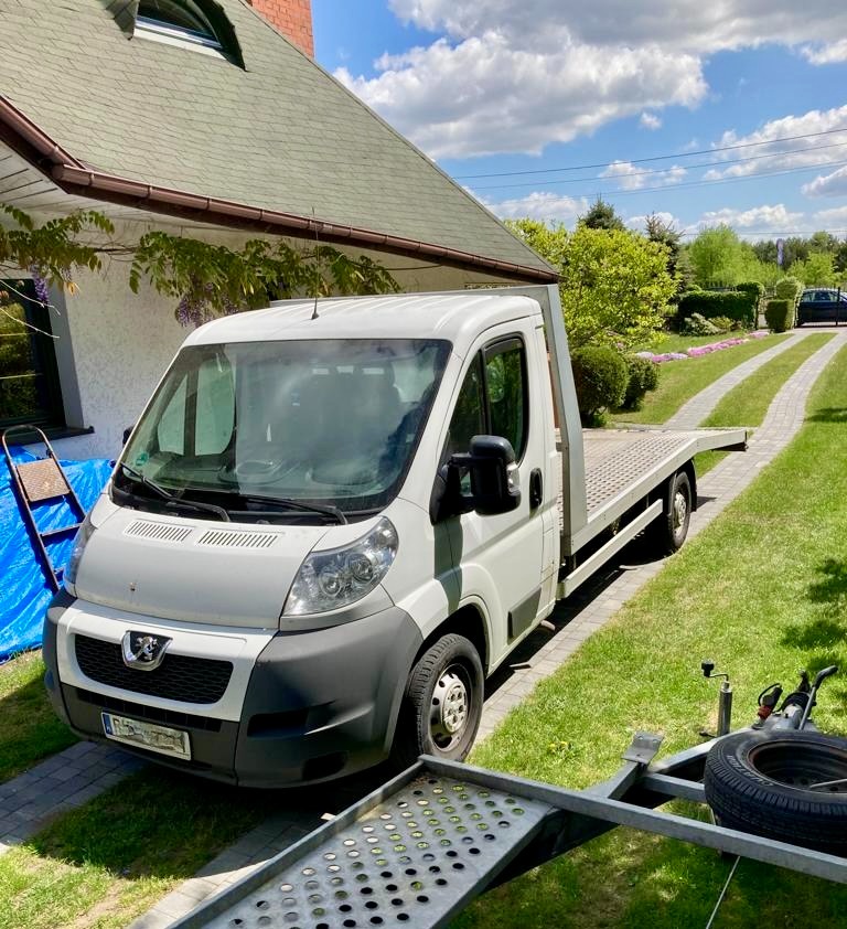Biała laweta Peugeot z platformą załadunkową zaparkowana na trawniku przed domem z zielonym dachem, obok przyczepka z kołem zapasowym.