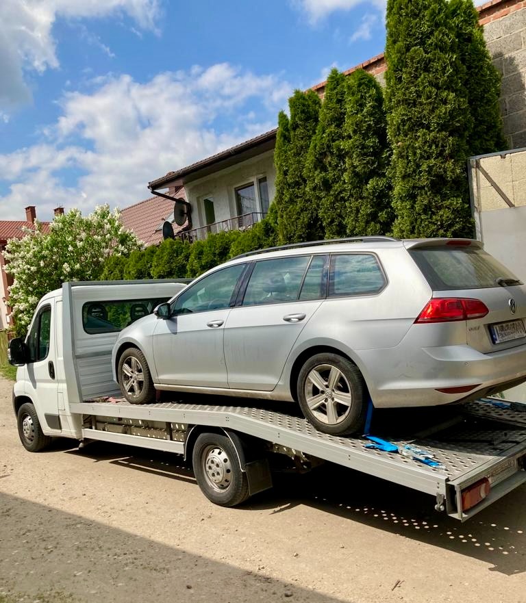 Srebrny samochód osobowy typu kombi na platformie lawety w trakcie załadunku, zabezpieczony niebieskimi pasami transportowymi, z widocznym fragmentem budynku mieszkalnego w tle.