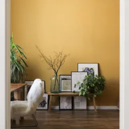Ściana pomalowana na kolor musztardowy w pokoju z drewnianą podłogą, dekorowana roślinami, szklanym wazonem z gałązkami i grafikami w czarnych ramkach, widok zza białych drzwi.