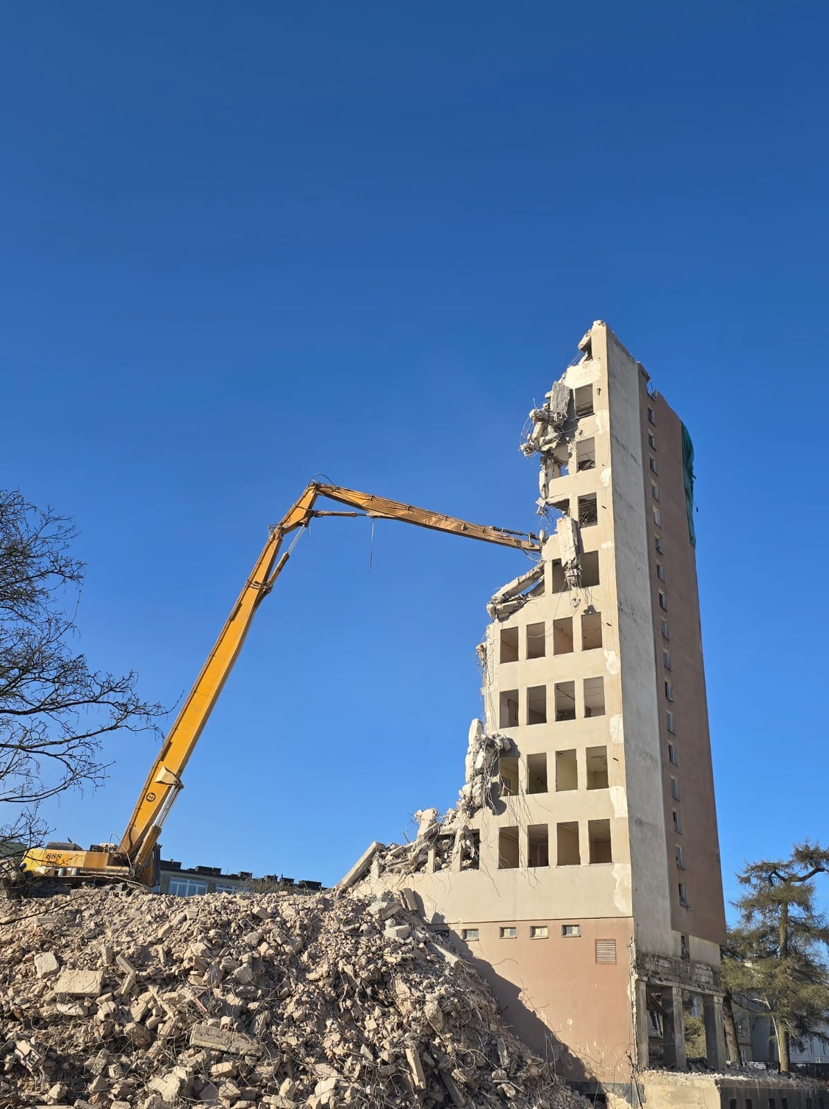 Żółty dźwig z długim ramieniem rozbiera wysoki budynek z odsłoniętymi oknami. Gruz leży u podnóża budynku na tle błękitnego nieba.