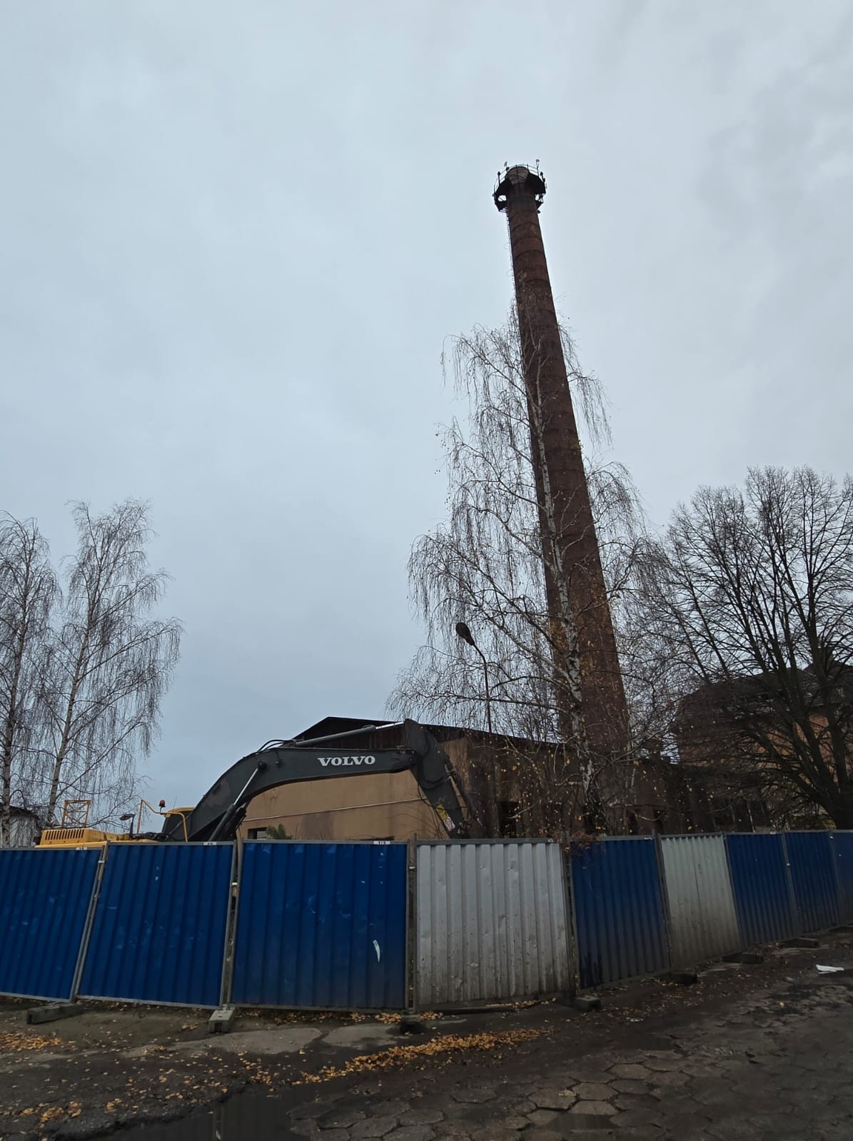 Żuraw Volvo na tle ceglanego komina i ogrodzenia. Rozbiórka budynku w pochmurny dzień. Widoczne drzewa bez liści.