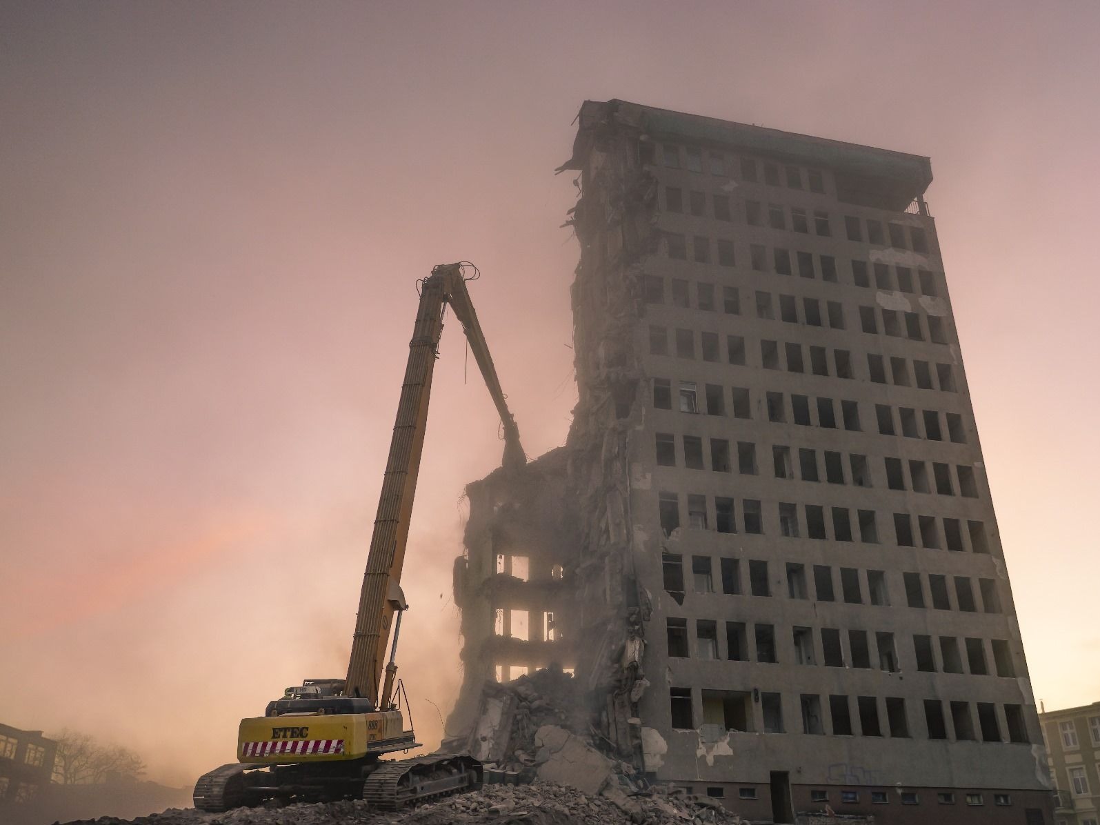 Żółty dźwig z długim ramieniem rozbiera wysoki, szary budynek w zapylonym otoczeniu. Widoczne gruzy i fragmenty konstrukcji. Ujęcie z dołu, w tle zamglone niebo.