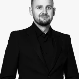 Portret mężczyzny w czarnym garniturze i koszuli, z lekkim uśmiechem, na białym tle. Zdjęcie czarno-białe.