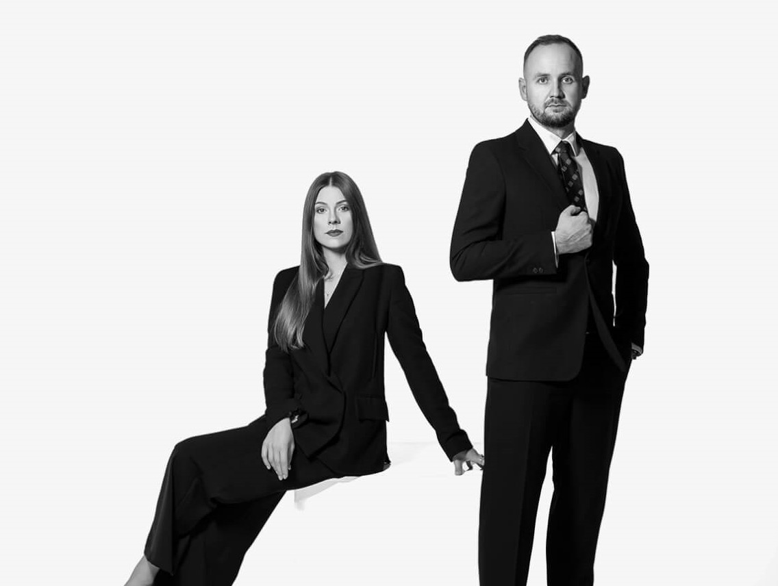 Portret biznesowy: Kobieta siedząca w garniturze i mężczyzna stojący w garniturze, oboje patrzą w obiektyw na białym tle.