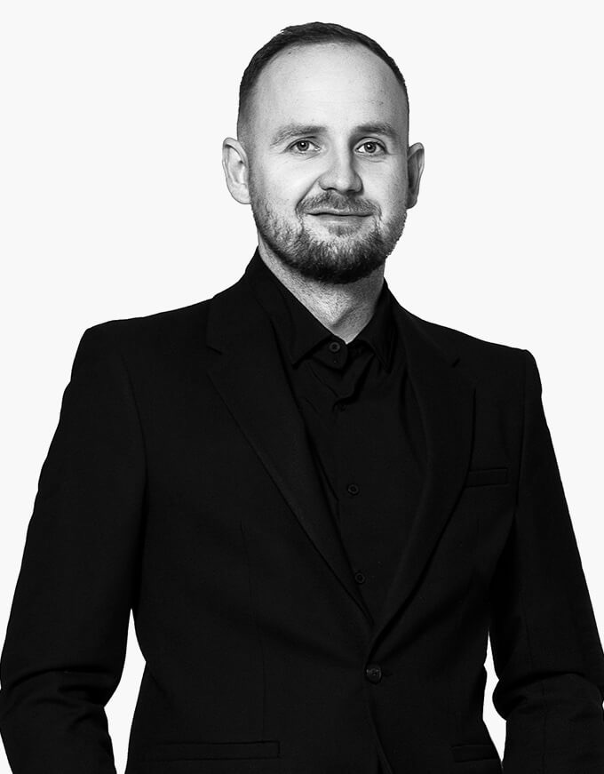 Portret mężczyzny w czarnym garniturze i koszuli, z lekkim uśmiechem, na białym tle. Zdjęcie czarno-białe.