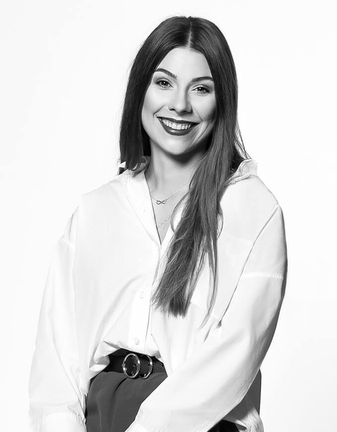 Portret uśmiechniętej kobiety w białej koszuli i ciemnej spódnicy, z naszyjnikiem nieskończoności, na jasnym tle.