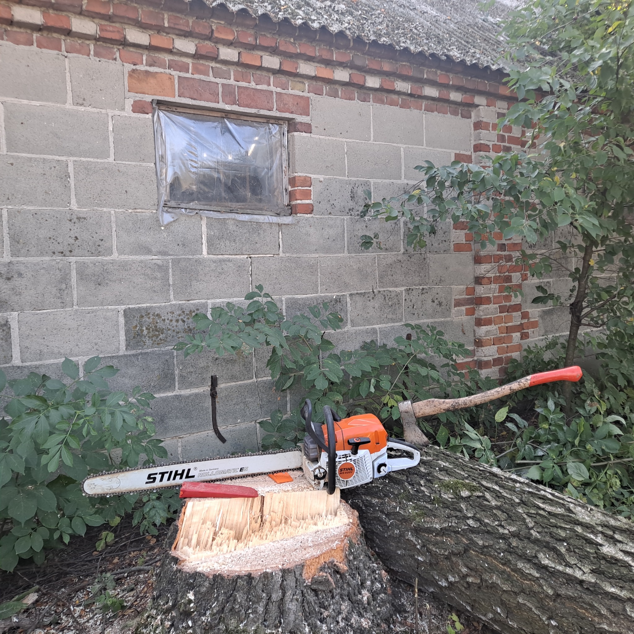 Pomarańczowo-biała piła spalinowa Stihl MS 400E i siekiera z czerwonym trzonkiem leżą na ściętym pniu drzewa przed budynkiem z szarego pustaka i czerwonej cegły, otoczone zielenią.
