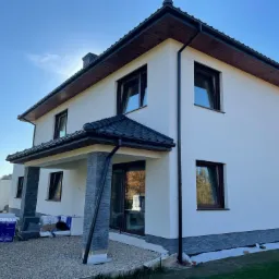 Nowoczesny dom jednorodzinny z jasną elewacją, ciemną dachówką i stolarką okienną, widoczne pakiety styropianu na posesji sugerujące prace wykończeniowe.